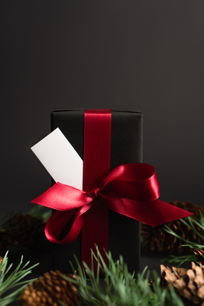 caja de regalo envuelta con cinta roja y tarjeta en blanco cerca de ramas de abeto borrosas y conos de pino en negro - Foto, imagen