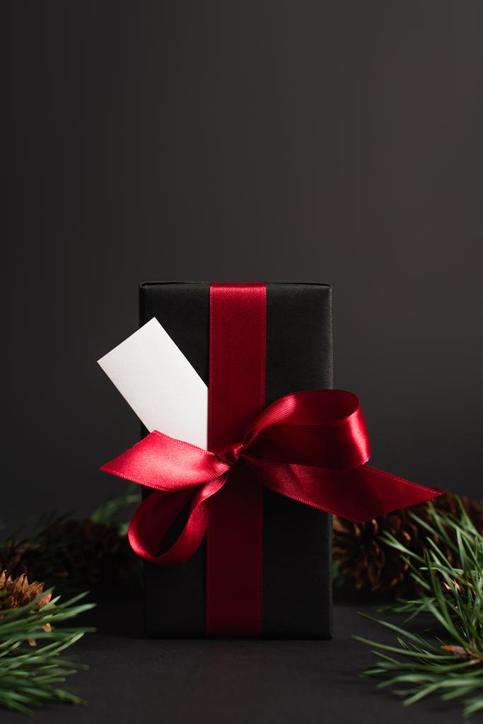 Geschenkpapier mit roter Schleife und leerer Karte in der Nähe verschwommener Tannenzweige auf schwarzem Grund - Foto, Bild