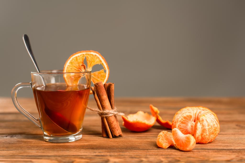 szklaną filiżankę herbaty w plasterkach pomarańczy w pobliżu kijów cynamonowych i obranej mandarynki wyizolowanej na szaro - Zdjęcie, obraz