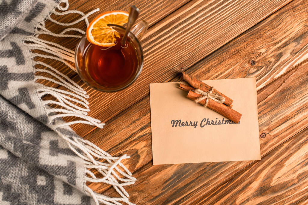 vista dall'alto di tazza di tè con affettata arancione vicino cannella bastoncini su biglietto di auguri con allegra scritta di Natale su superficie di legno  - Foto, immagini
