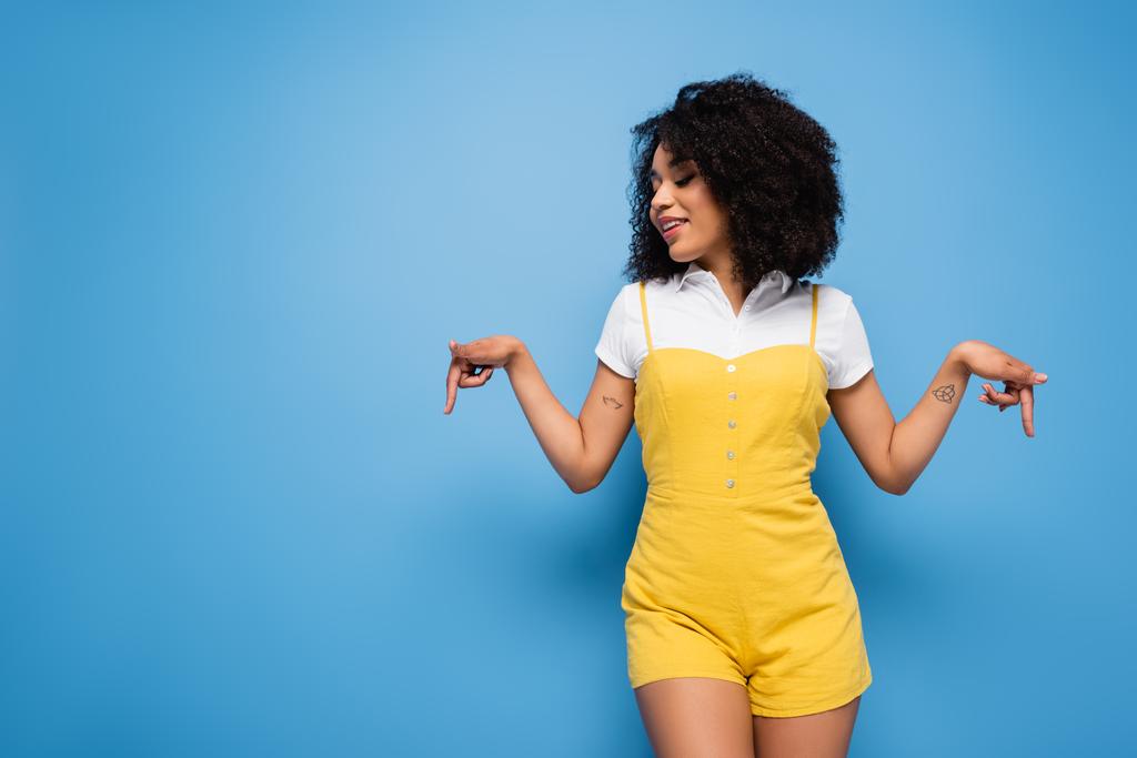 smiling african american woman in trendy jumpsuit pointing down with fingers on blue - Photo, Image