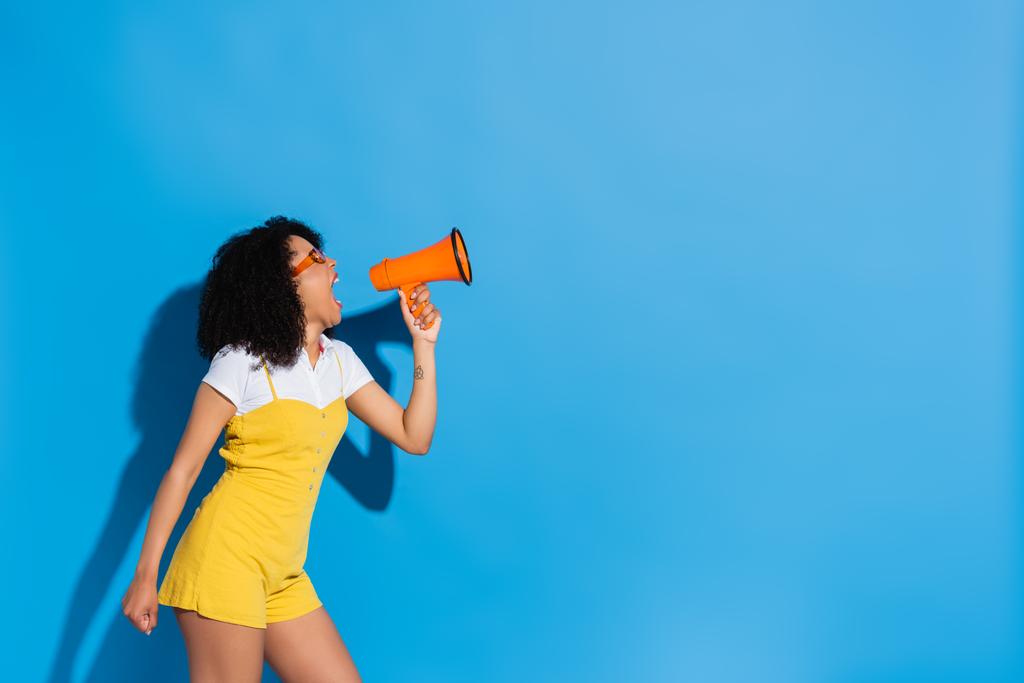Afričanky americká žena ve žluté kombinéze křičí v megafonu na modré - Fotografie, Obrázek