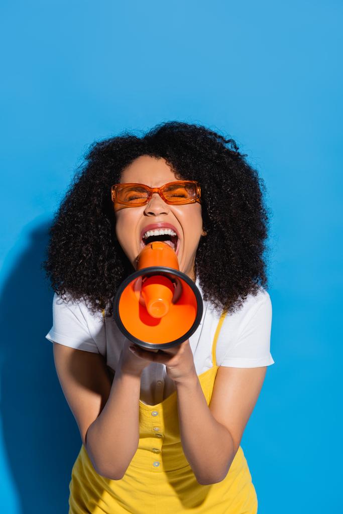 Ενθουσιασμένη Αφροαμερικανή γυναίκα με πορτοκαλί γυαλιά ουρλιάζοντας σε μεγάφωνο στο μπλε - Φωτογραφία, εικόνα