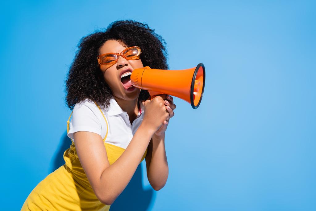 agitata donna afroamericana in occhiali alla moda urlando in megafono su blu - Foto, immagini