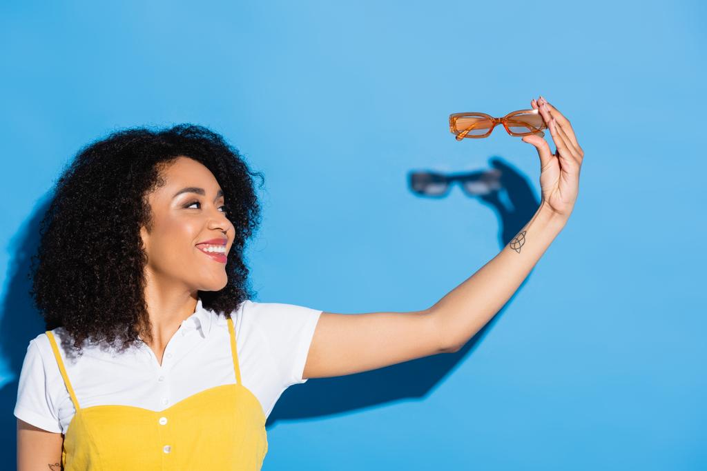 femme américaine africaine gaie tenant des lunettes de vue à la mode sur bleu - Photo, image