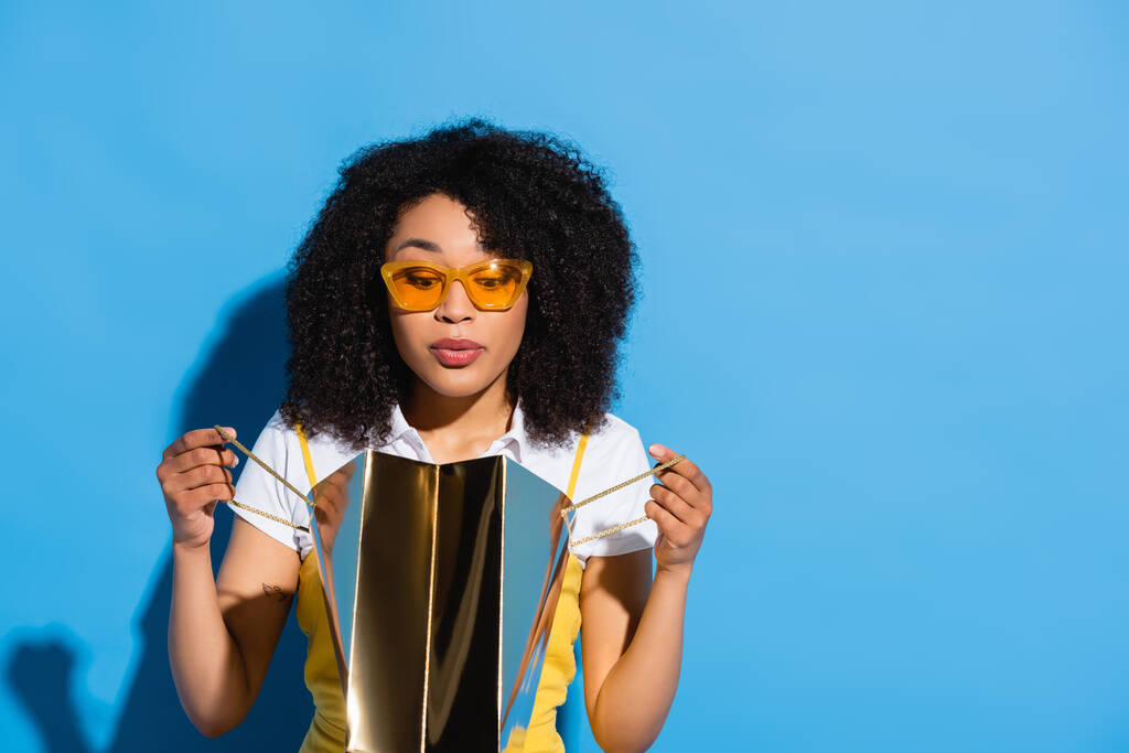überraschte Afroamerikanerin mit gelber Brille blickt in glänzende Einkaufstasche auf blauem Grund - Foto, Bild