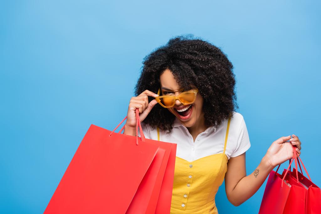 mujer afroamericana emocionada con bolsas de compras tocando gafas de moda aisladas en azul - Foto, Imagen
