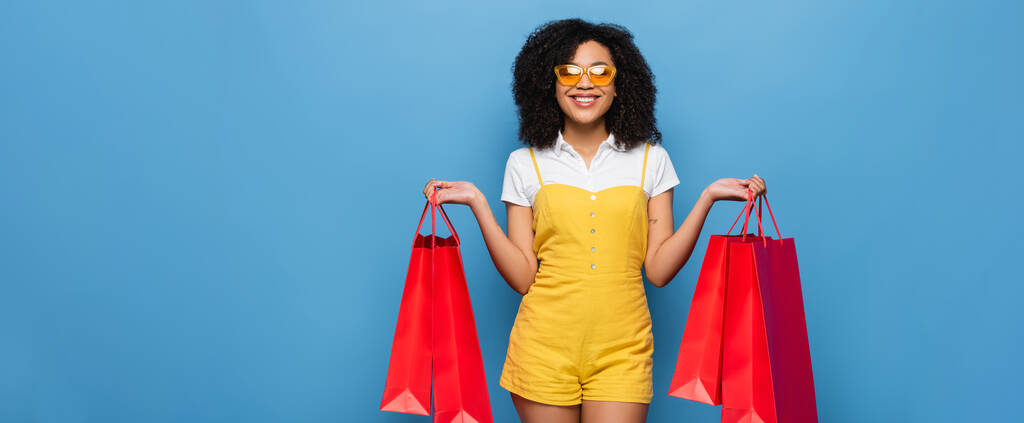 vidám afro-amerikai nő sárga kezeslábasban és szemüvegben, kék zászlón elszigetelt bevásárlószatyrokkal. - Fotó, kép