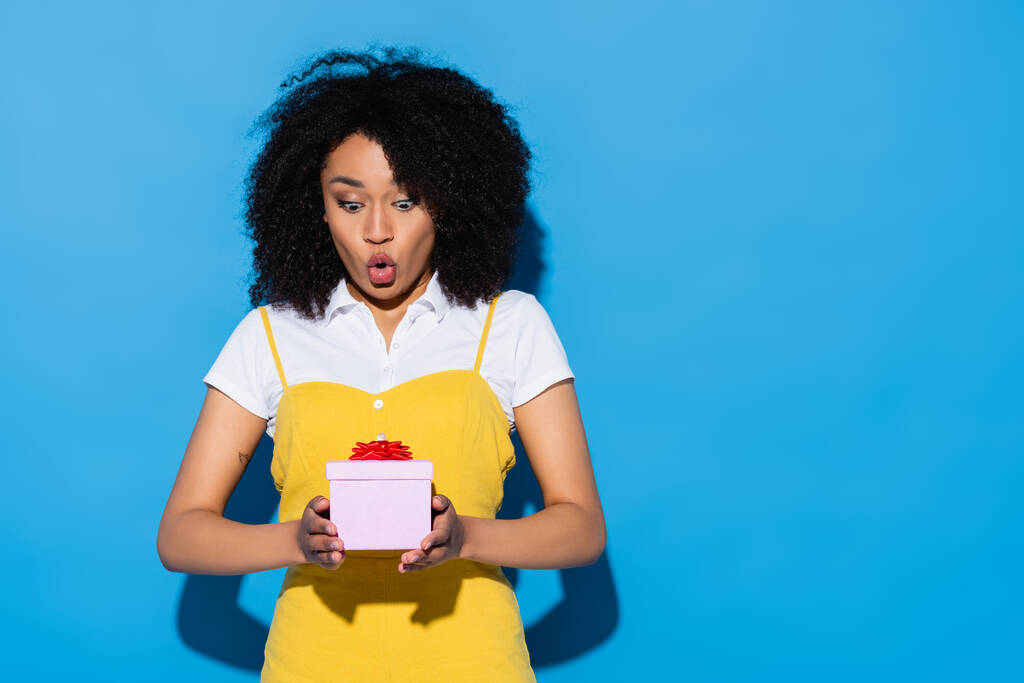 mujer afroamericana asombrada con boca abierta sosteniendo caja de regalo en azul - Foto, Imagen