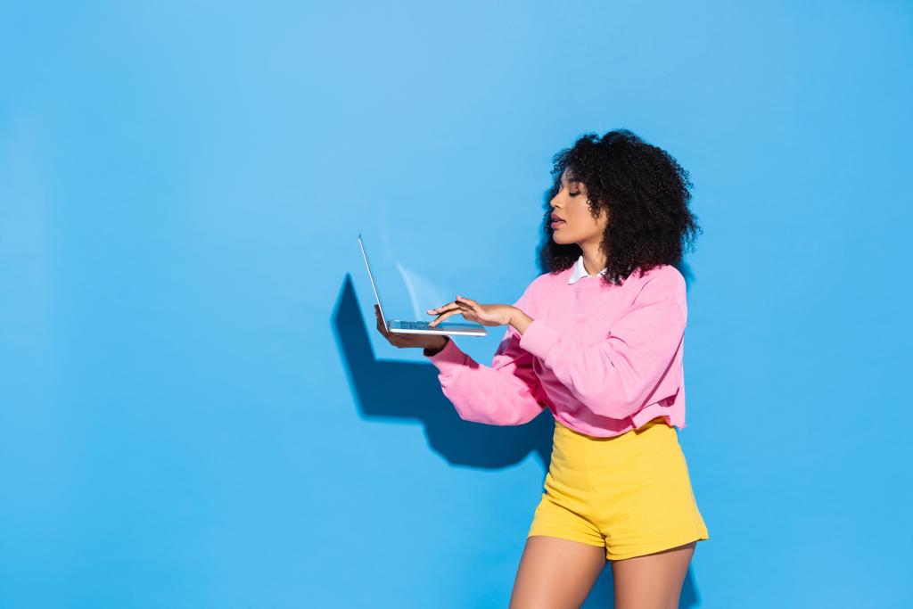 mujer afroamericana joven en pantalones cortos amarillos y sudadera rosa usando el ordenador portátil en azul - Foto, imagen
