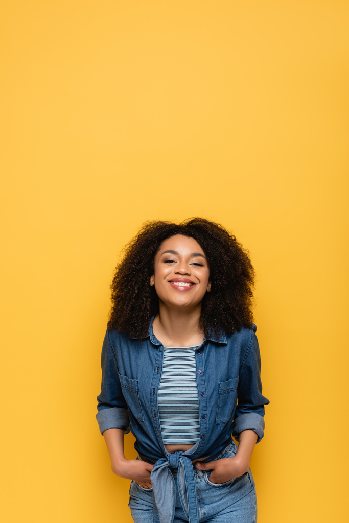 счастливая африканская американка смотрит в камеру, стоя с руками в карманах, изолированных на желтом - Фото, изображение