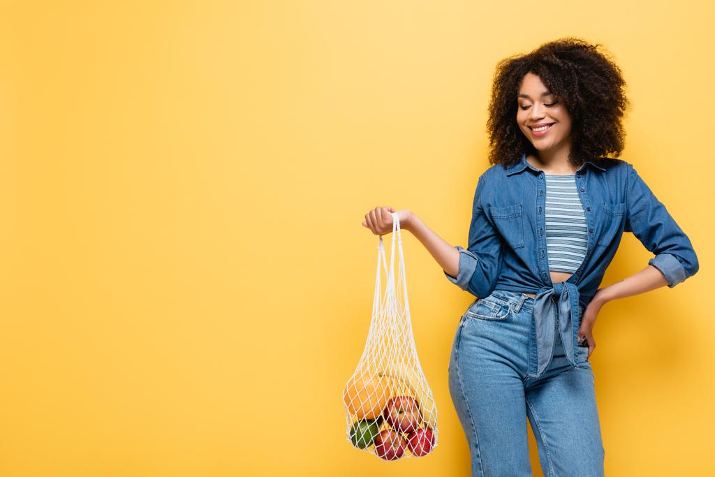 alegre mujer afroamericana sosteniendo bolsa de cuerda con frutas frescas mientras posa con la mano en la cadera en amarillo - Foto, Imagen