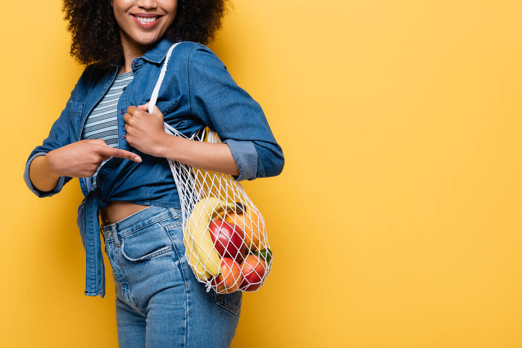 abgeschnittene Ansicht einer lächelnden Afroamerikanerin, die auf eine Plastiktüte mit reifen Früchten zeigt, vereinzelt auf gelb - Foto, Bild