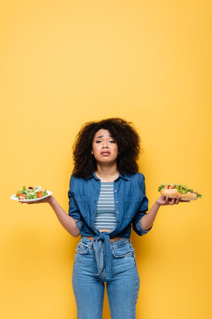 Sebze salatası ve sosisli sandviçli üzgün Afrikalı Amerikalı kadın sarıya izole edilmiş kameraya bakıyor. - Fotoğraf, Görsel