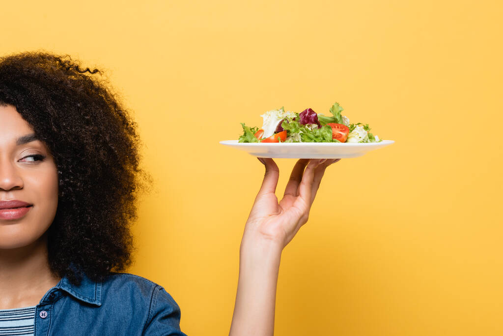 частичный вид африканской американки, держащей тарелку со свежим салатом, изолированным на желтом - Фото, изображение