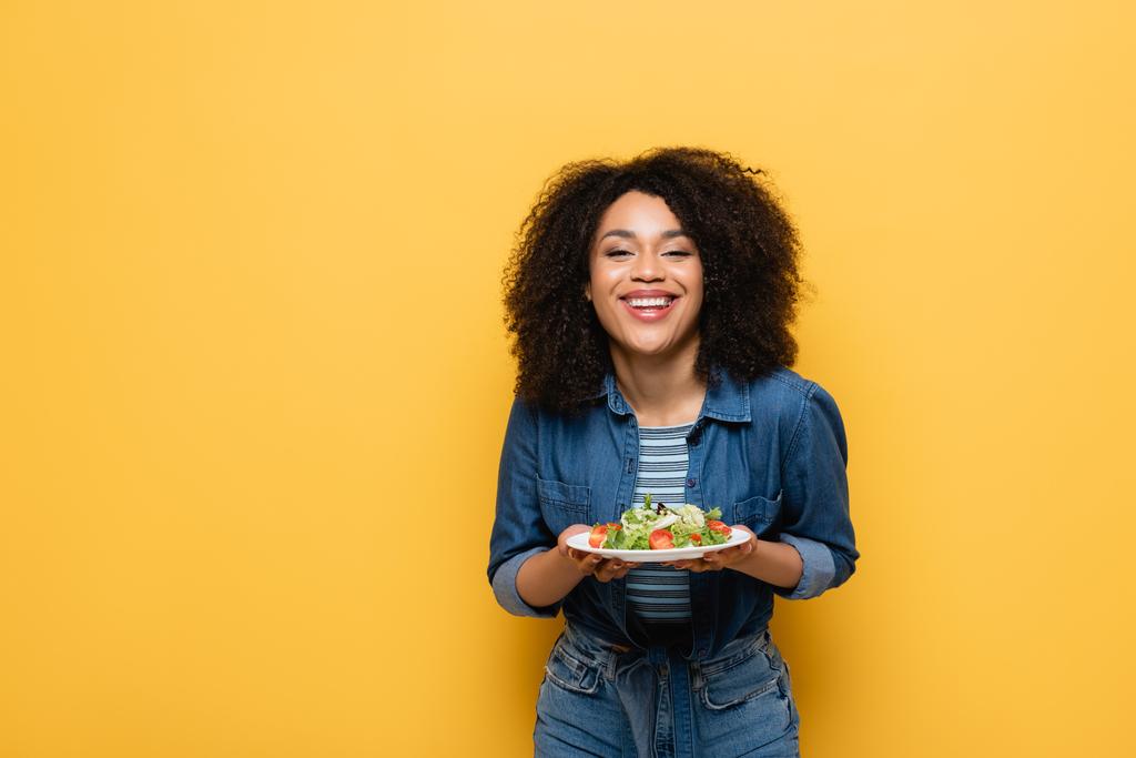 happy african american woman holding plate with fresh vegetable salad isolated on yellow - Photo, Image