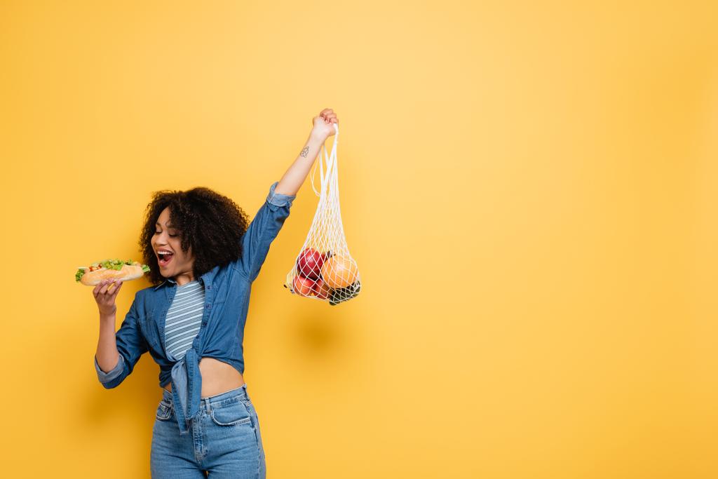 アフリカ系アメリカ人の女性がホットドッグをかむ一方、黄色に手を挙げて新鮮な果物でストリングバッグを保持 - 写真・画像