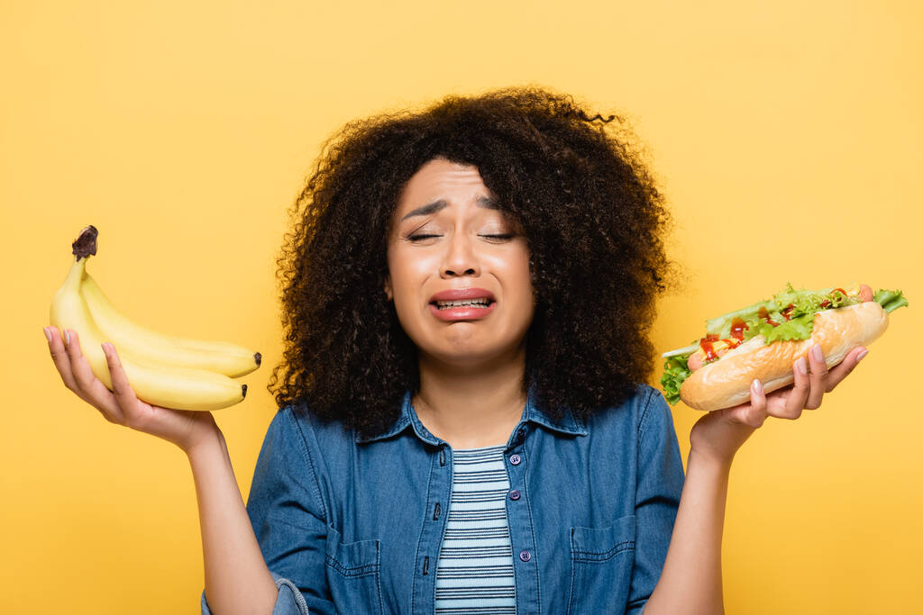 upset african american woman choosing between fresh bananas and hot dog isolated on yellow - Photo, Image