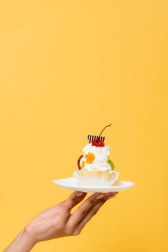 partial view of african american woman holding delicious cupcake with whipped cream isolated on yellow - Photo, Image