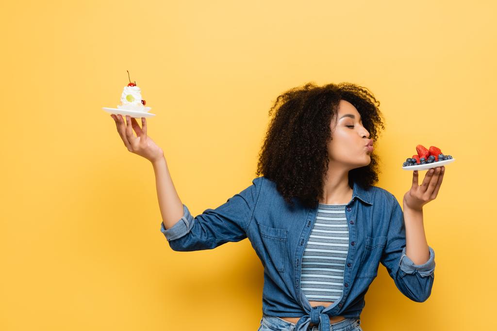 donna afro-americana pouting labbra vicino a bacche fresche, mentre tenendo cupcake su giallo - Foto, immagini