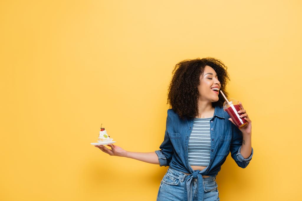 happy african american woman holding cupcake while drinking fresh smoothie on yellow - Photo, Image