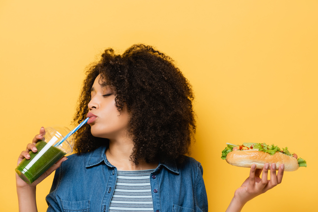 african american woman with closed eyes drinking fresh smoothie while holding hot dog isolated on yellow - Photo, Image