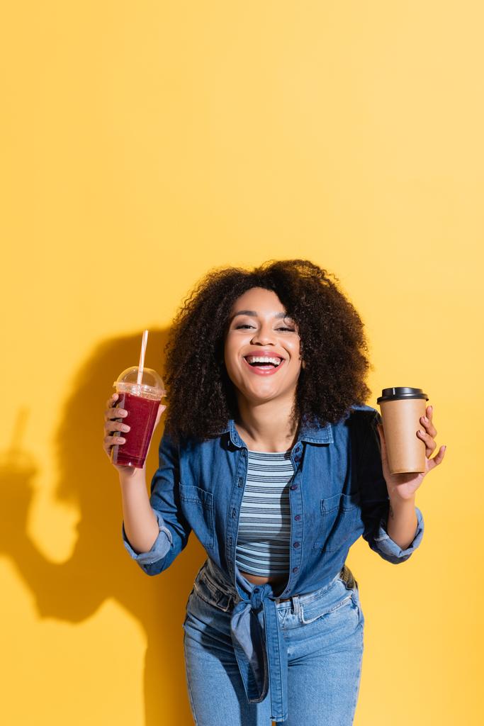 fröhliche afrikanisch-amerikanische Frau mit Kaffee und frischem Smoothie lächelt in die Kamera auf gelb - Foto, Bild