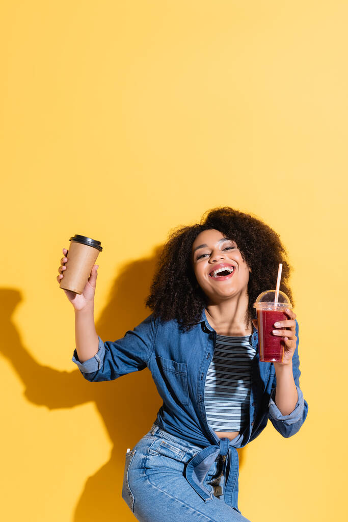 ενθουσιασμένοι αφροαμερικανός γυναίκα με smoothie και καφέ για να πάει κοιτάζοντας κάμερα σε κίτρινο - Φωτογραφία, εικόνα