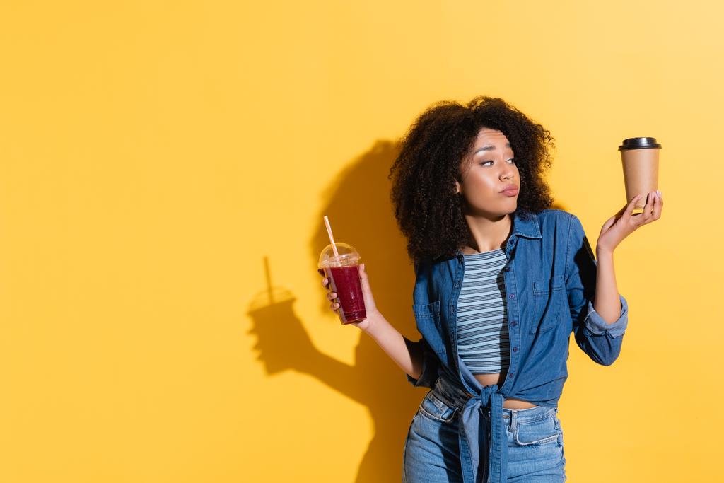 discouraged african american woman holding fresh smoothie while looking at coffee to go on yellow - Photo, Image