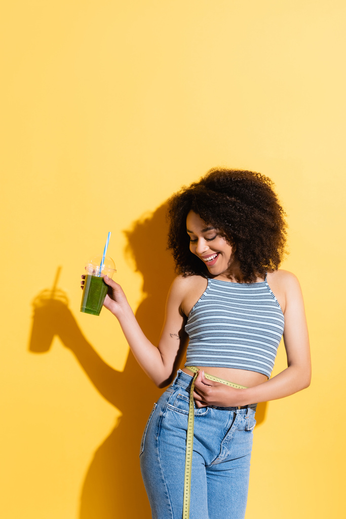 alegre afroamericana mujer medición cintura mientras celebración fresco smoothie en amarillo - Foto, Imagen