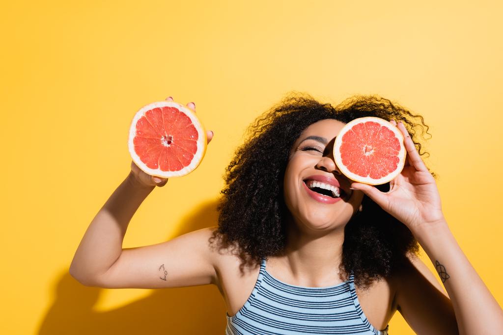 aufgeregte afrikanisch-amerikanische Frau lacht, während sie Hälften frischer Grapefruit auf gelb hält - Foto, Bild