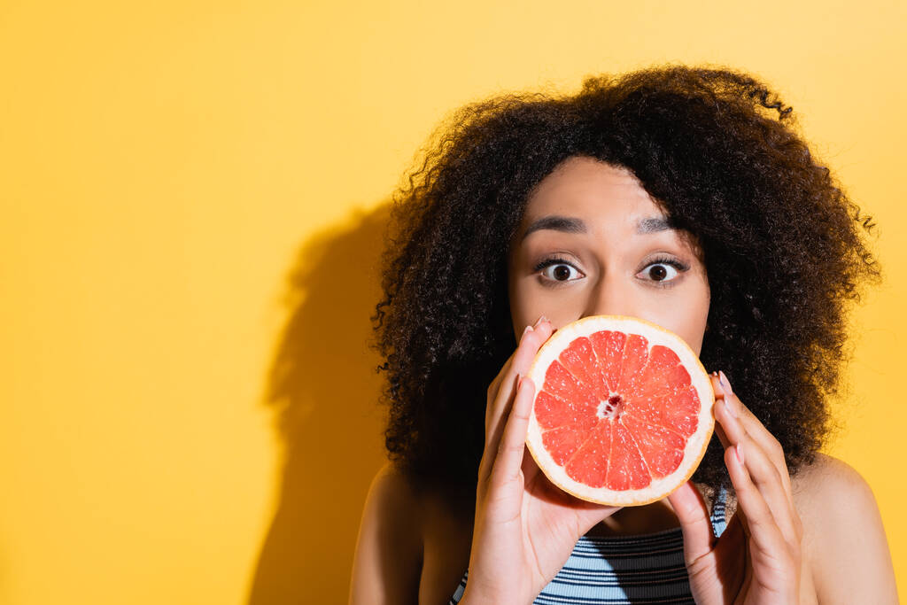 mladá africká americká žena zakrývající ústa s polovinou čerstvého grapefruitu na žluté - Fotografie, Obrázek
