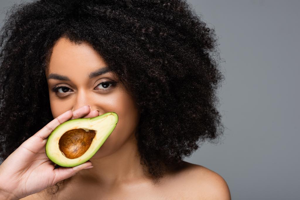 hübsche afrikanisch-amerikanische Frau hält die Hälfte der frischen Avocado in Gesichtsnähe isoliert auf grau - Foto, Bild
