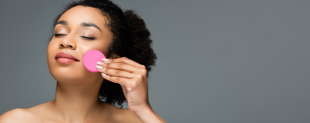 donna afroamericana ad occhi chiusi che applica fondotinta viso con spugna cosmetica isolata su grigio, banner - Foto, immagini