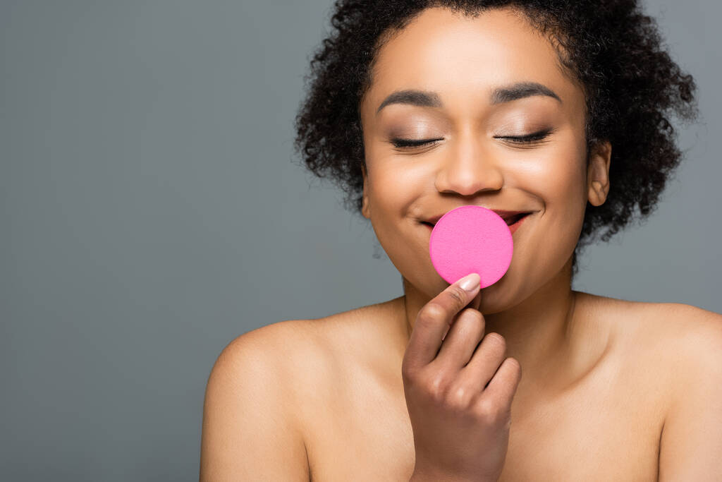 cheerful african american woman with closed eyes and naked shoulders holding cosmetic sponge near lips isolated on grey - Photo, Image