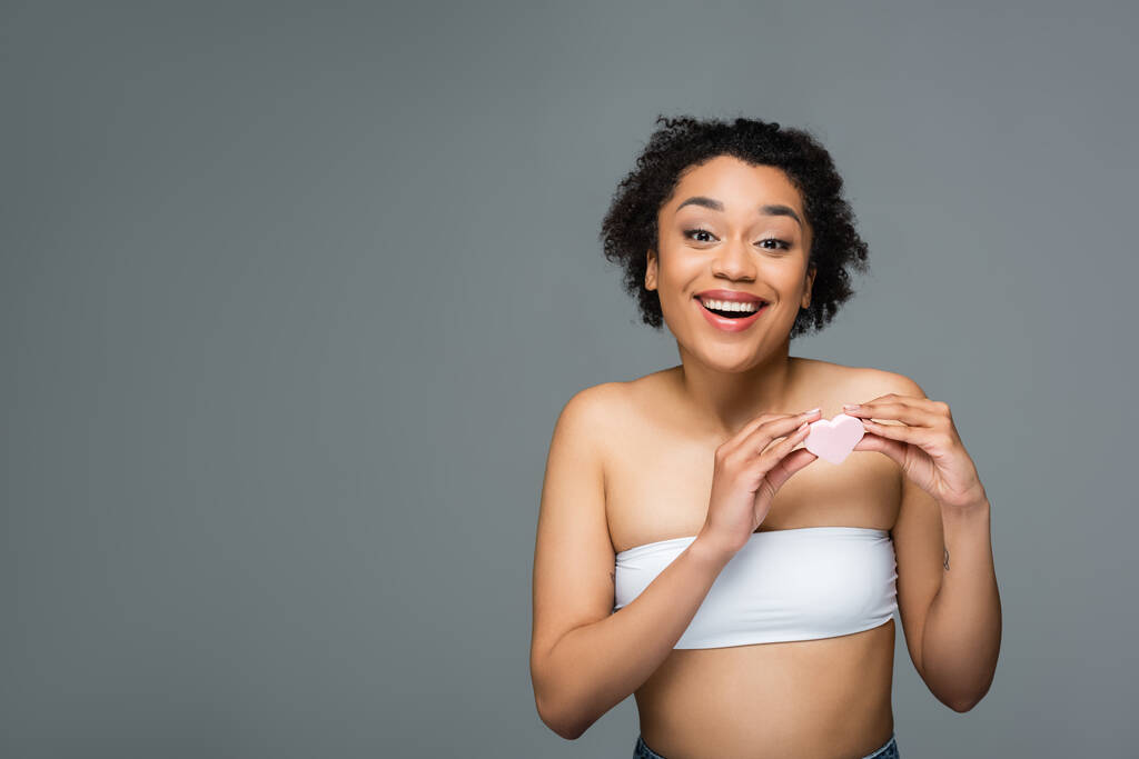 excitada mujer afroamericana mirando a la cámara mientras sostiene una esponja cosmética en forma de corazón aislada en gris - Foto, Imagen