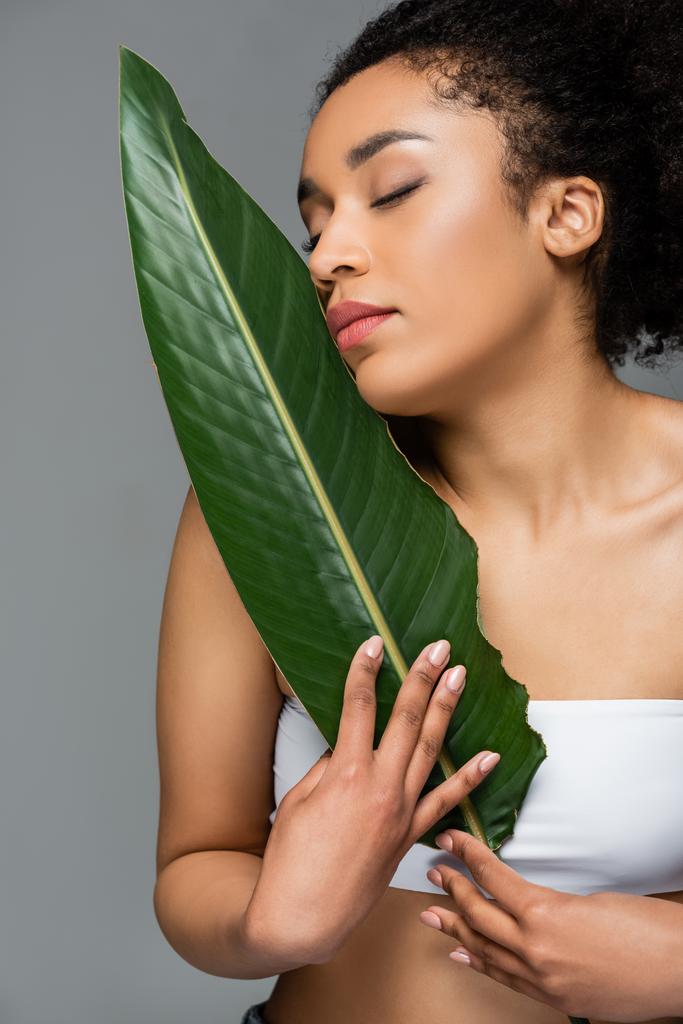 femme afro-américaine avec une peau parfaite et les yeux fermés posant avec une feuille verte isolée sur gris - Photo, image