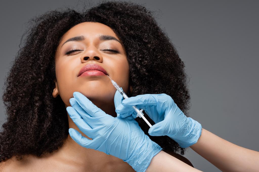 cosmetoloog in latex handschoenen doen schoonheid injectie naar lippen van Afrikaans amerikaanse vrouw geïsoleerd op grijs - Foto, afbeelding