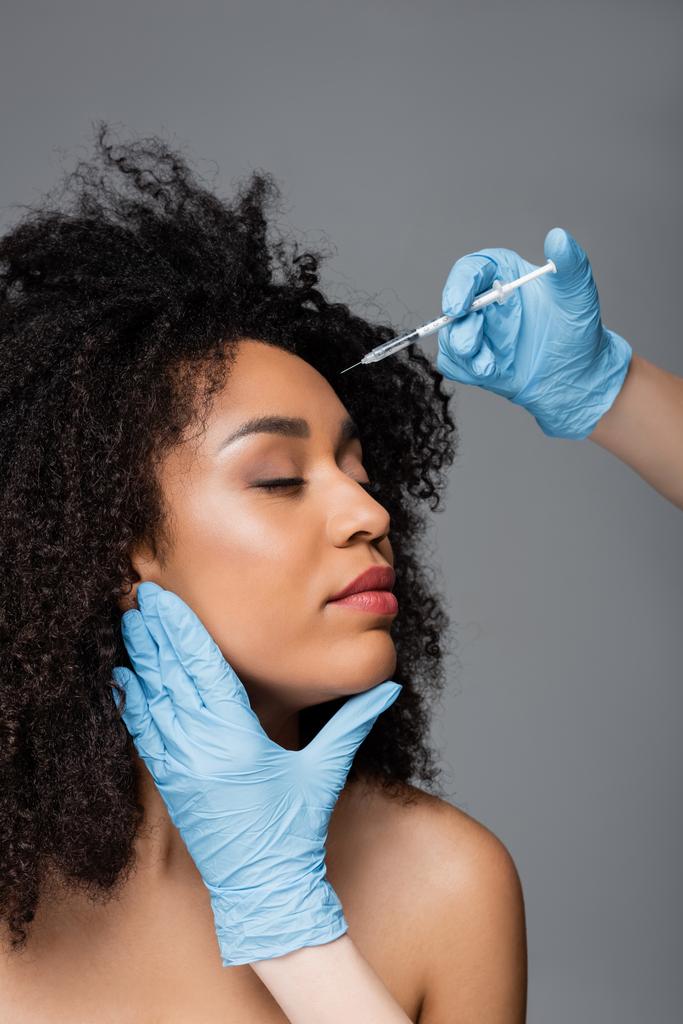 kosmetolog w lateksowych rękawiczkach robi zastrzyk urody afrykańskiej kobiecie wyizolowanej na szaro - Zdjęcie, obraz