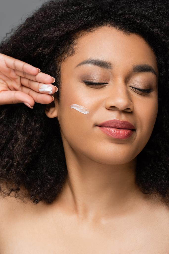 pleased african american woman with cosmetic cream on face posing with closed eyes isolated on grey - Photo, Image
