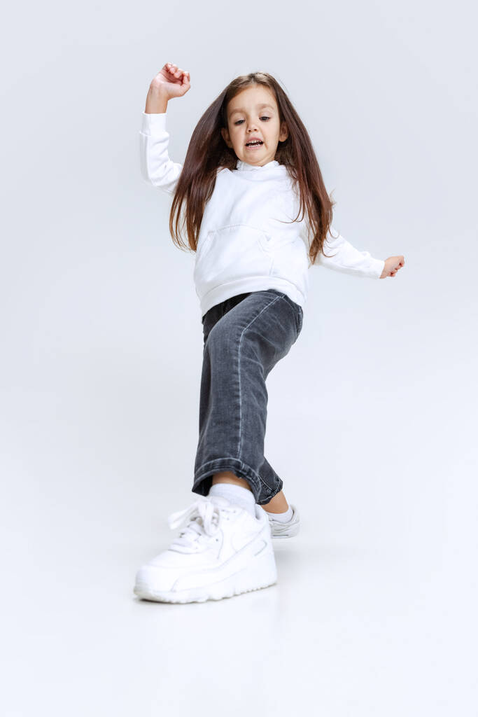 Full-length portret van klein schattig meisje, kind dragen hoodie met jeans stappen, sprongen geïsoleerd over witte studio achtergrond. - Foto, afbeelding