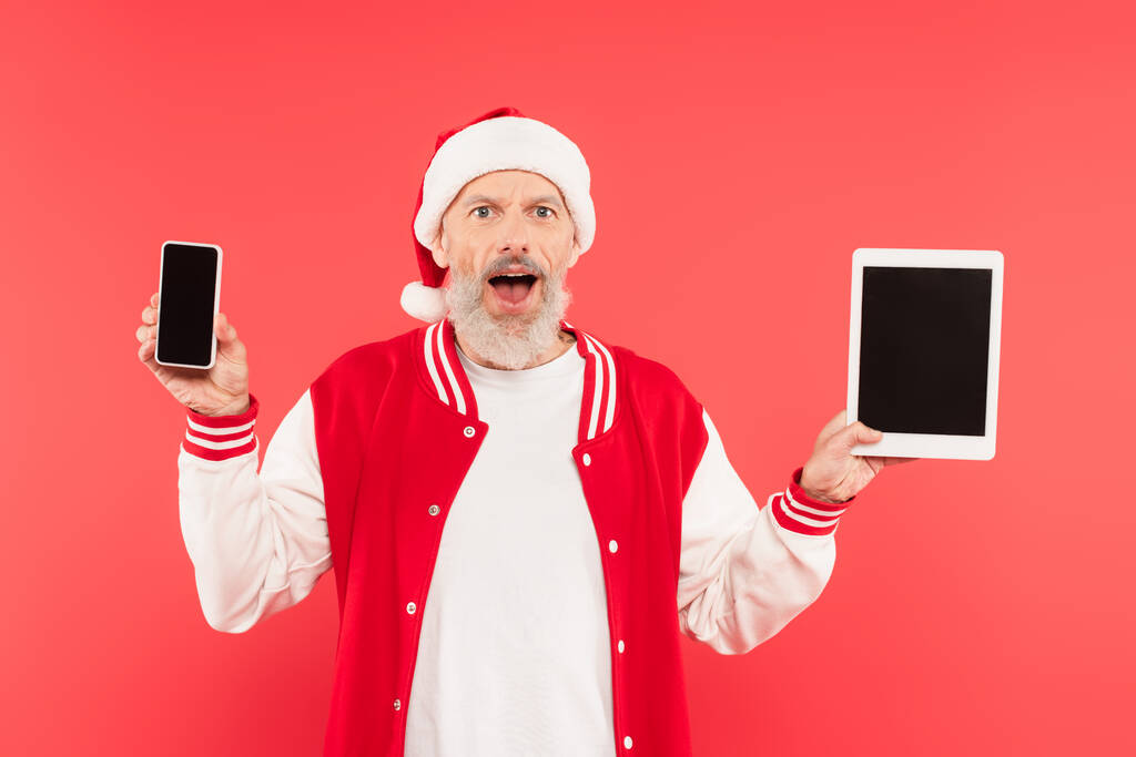 здивований чоловік середнього віку в капелюсі Санта тримає гаджети з порожнім екраном ізольовано на червоному
 - Фото, зображення