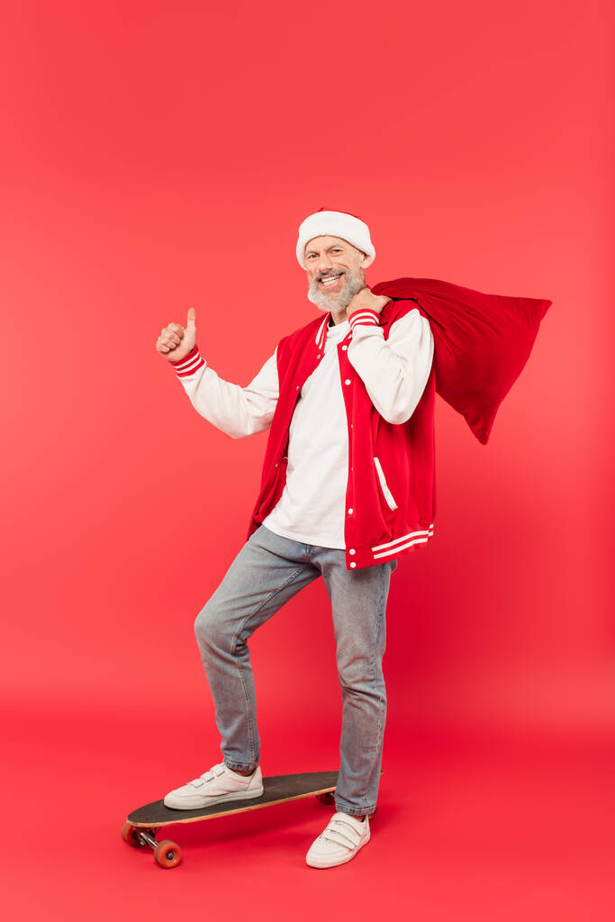 longitud completa de alegre hombre de mediana edad en sombrero sosteniendo santa saco mientras monta monopatín y mostrando el pulgar hacia arriba en rojo - Foto, Imagen