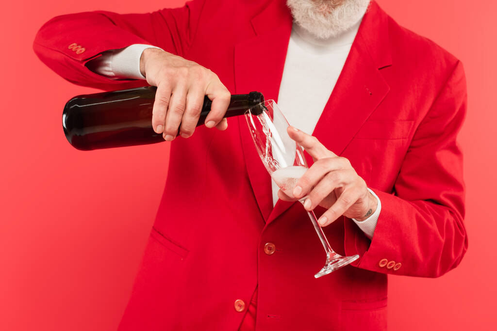 обрезанный вид бородатого мужчины средних лет в костюме, держащего бутылку, разливая шампанское в стакан, изолированный на красный - Фото, изображение