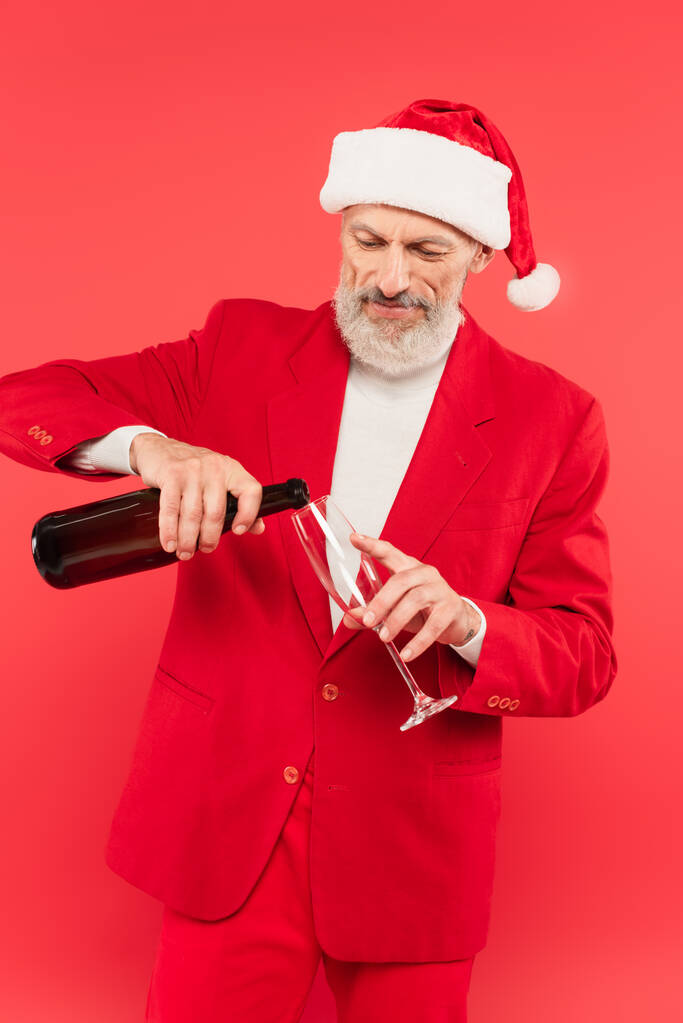 bearded middle aged man in suit and santa hat holding bottle while pouring champagne in glass isolated on red - Photo, Image