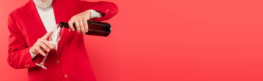 vista ritagliata dell'uomo barbuto di mezza età in tuta che tiene in bottiglia mentre versa champagne in vetro isolato sul rosso, banner - Foto, immagini
