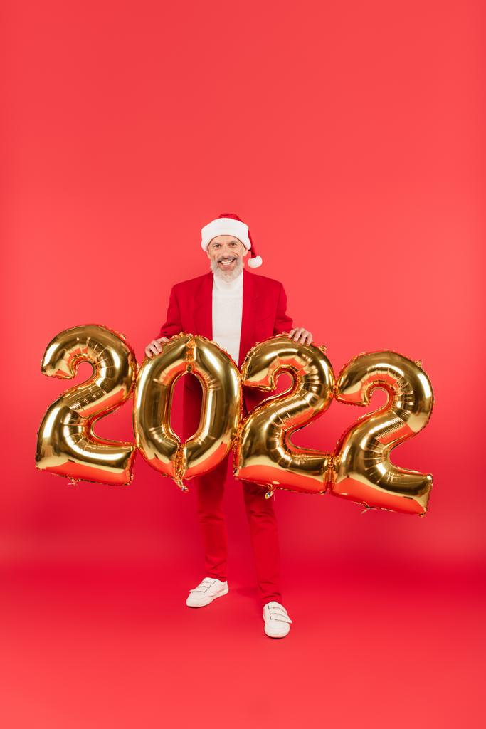 щасливий зрілий чоловік у капелюсі Санта стоїть біля 2022 повітряних кульок на червоному
 - Фото, зображення