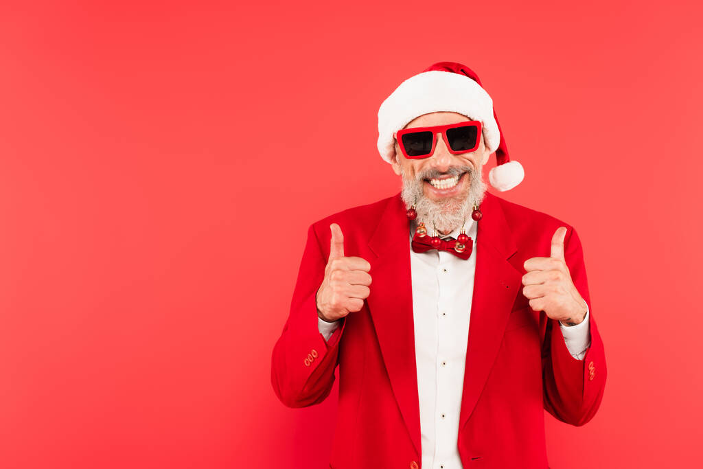 happy mature man in santa hat and sunglasses with christmas balls on beard showing thumbs up isolated on red - Photo, Image