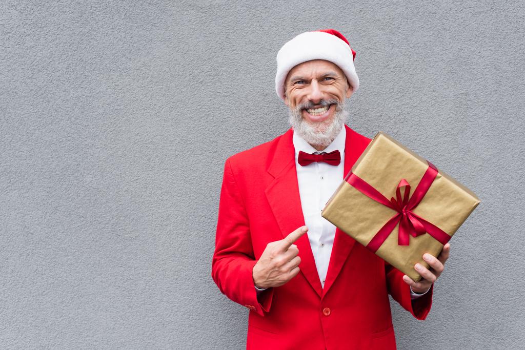 felice uomo maturo in cappello di Babbo Natale che punta al regalo avvolto vicino al muro grigio - Foto, immagini