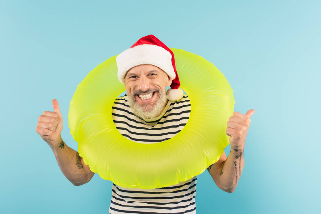 homme d'âge moyen positif dans le chapeau santa tenant anneau gonflable tout en montrant pouces isolés sur bleu - Photo, image